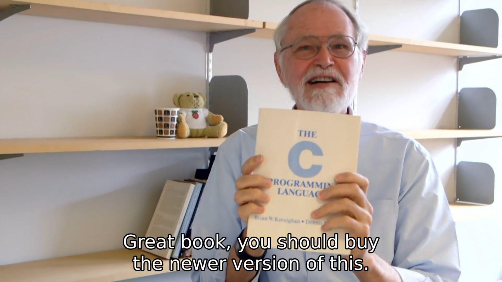 Brian Kernighan holds up the book "The C Programming Language" by Brian W. Kernighan and Dennis M. Ritchie. The subtitle says "Great book, you should buy the newer version of this."
